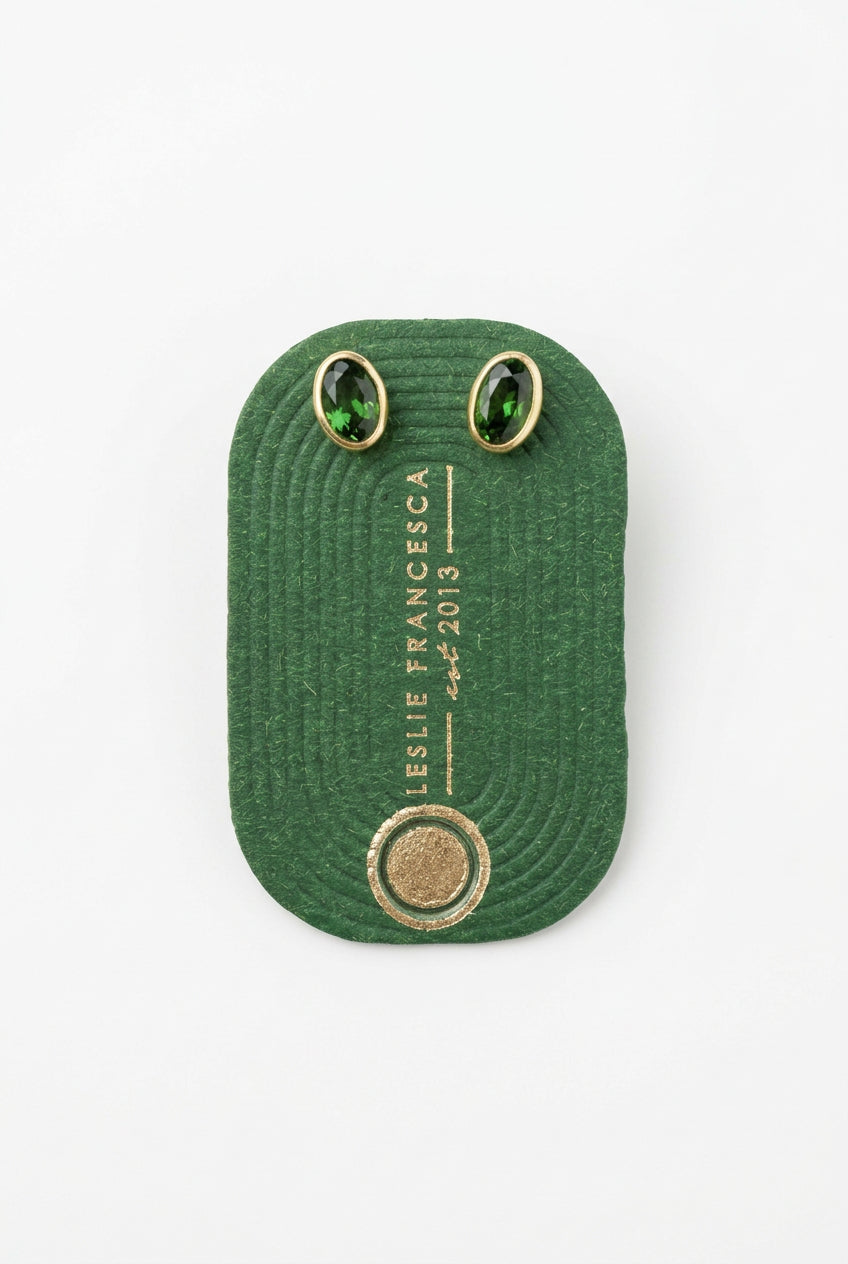 A dark green textured fabric patch with white printed text reading 'LESLIE FRANCESCA' and 'EST. 2013', featuring two oval green stone settings on a light wood background.