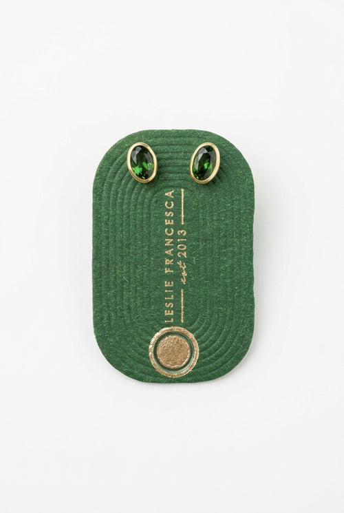 A dark green textured fabric patch with white printed text reading 'LESLIE FRANCESCA' and 'EST. 2013', featuring two oval green stone settings on a light wood background.